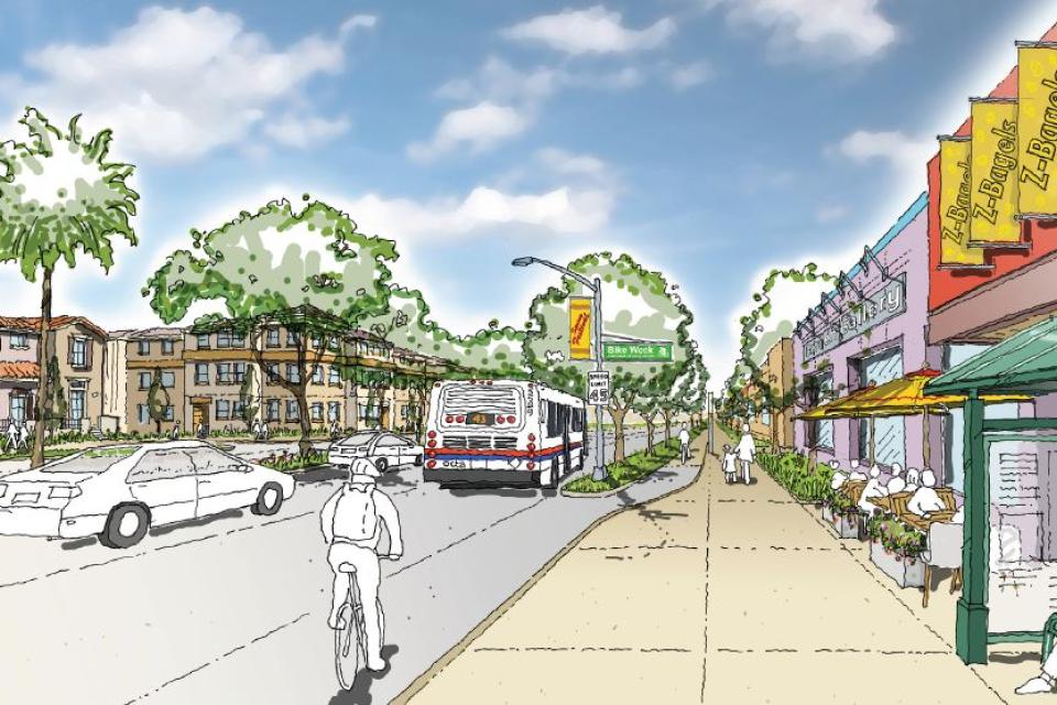 Street with shops, trees, cars, and a bus under a partly cloudy sky.